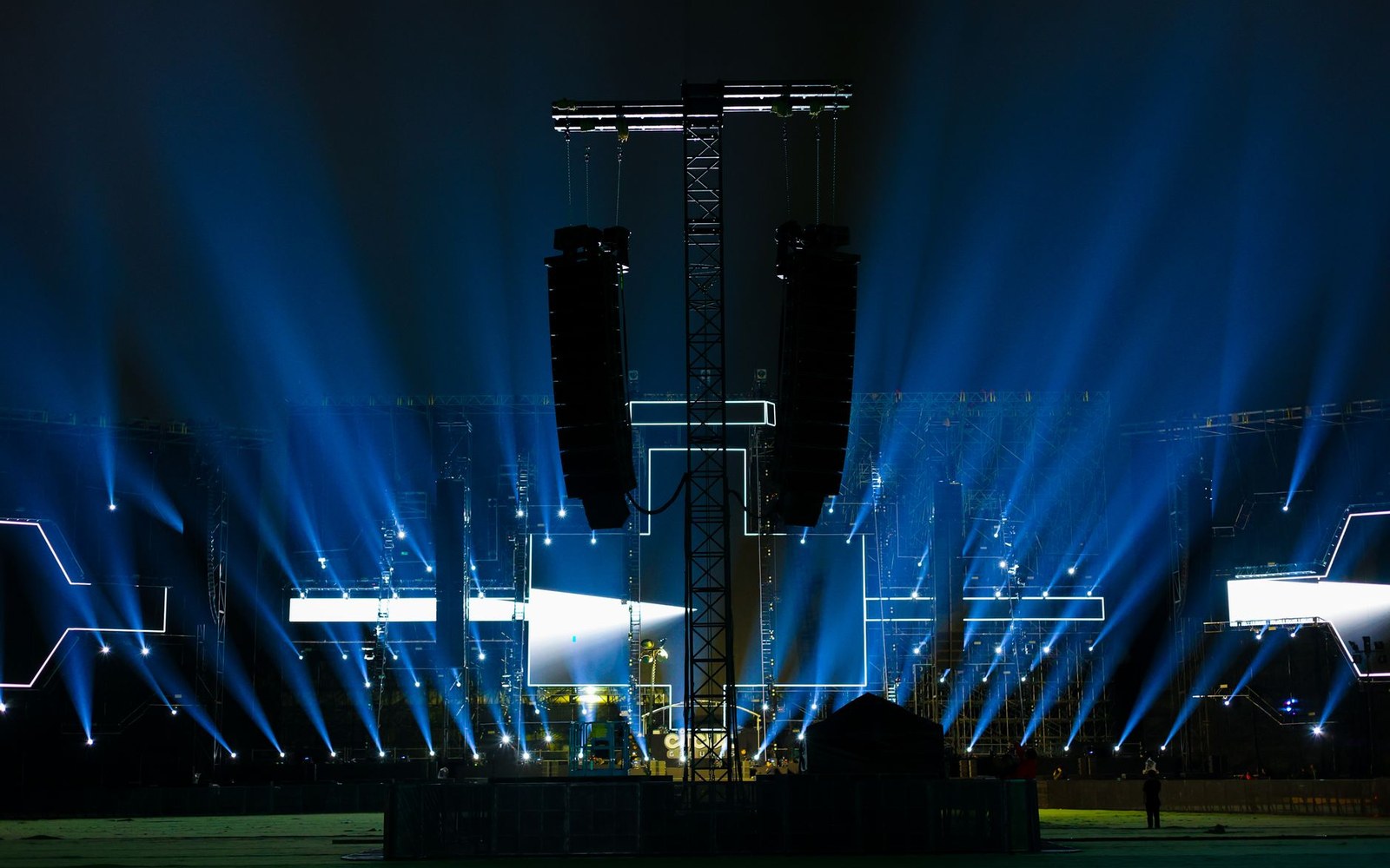 Blue stage lights beam outward from a concert rig with hanging line array speakers at night