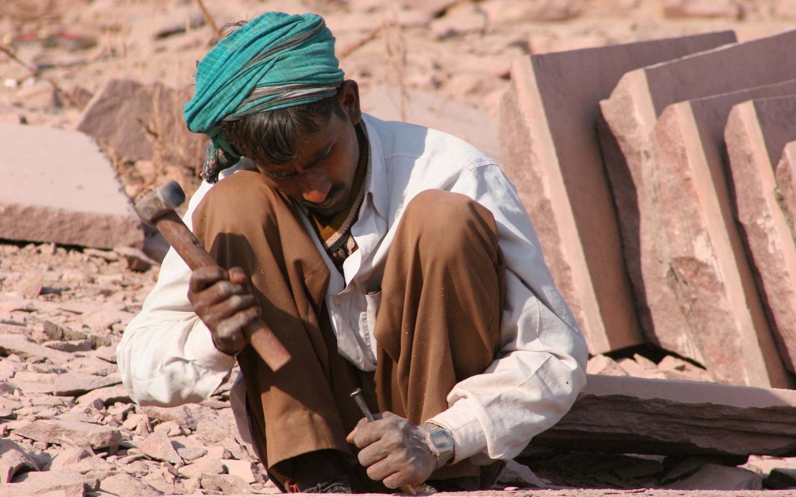 A turbaned worker crouches on rocky ground, chiseling a stone slab with hammer and chisel