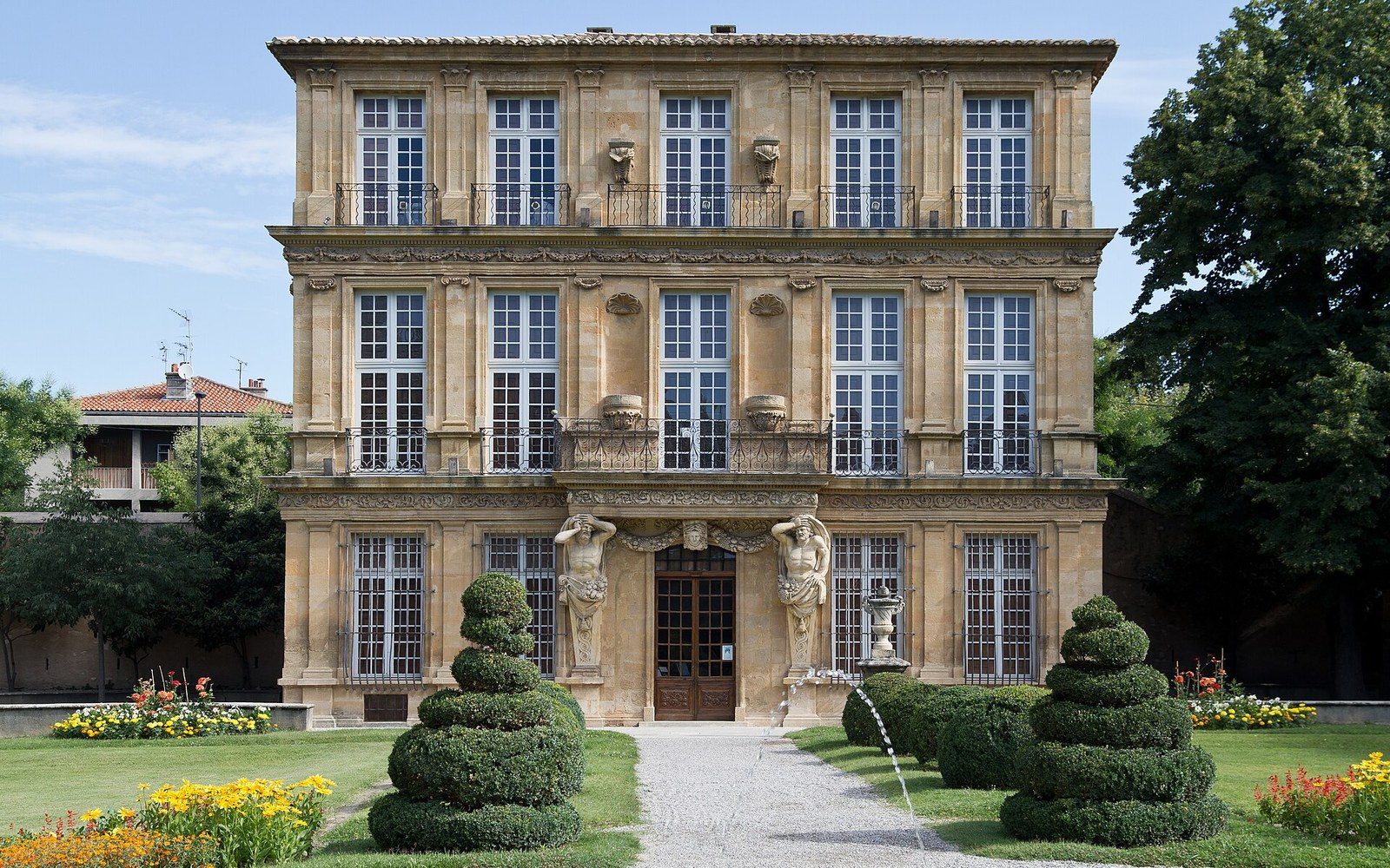 Three-story stone mansion with carved atlas figures flanking the entrance and manicured spiral topiaries