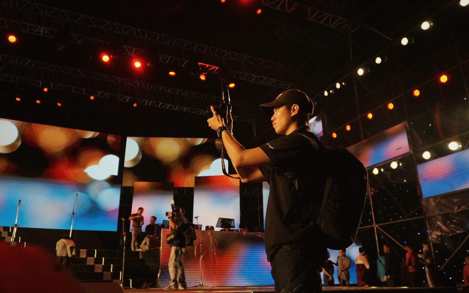 A man wearing a backpack films with a camera near a stage under rigging lights